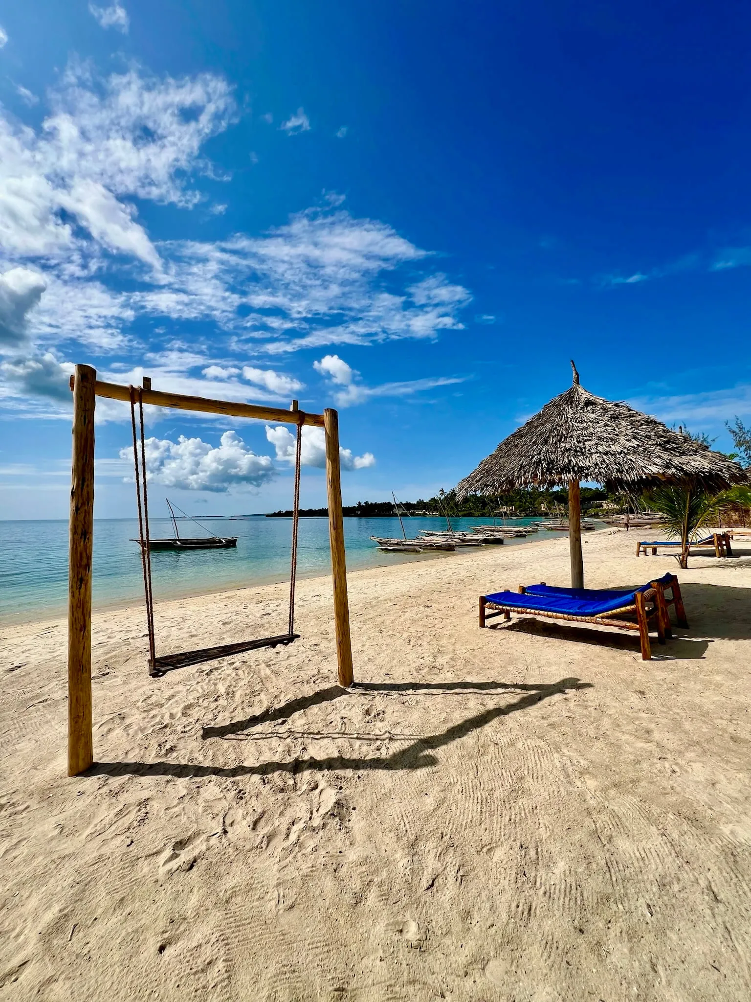 A wooden swing standing on the beach beside loungers and a thatched umbrella