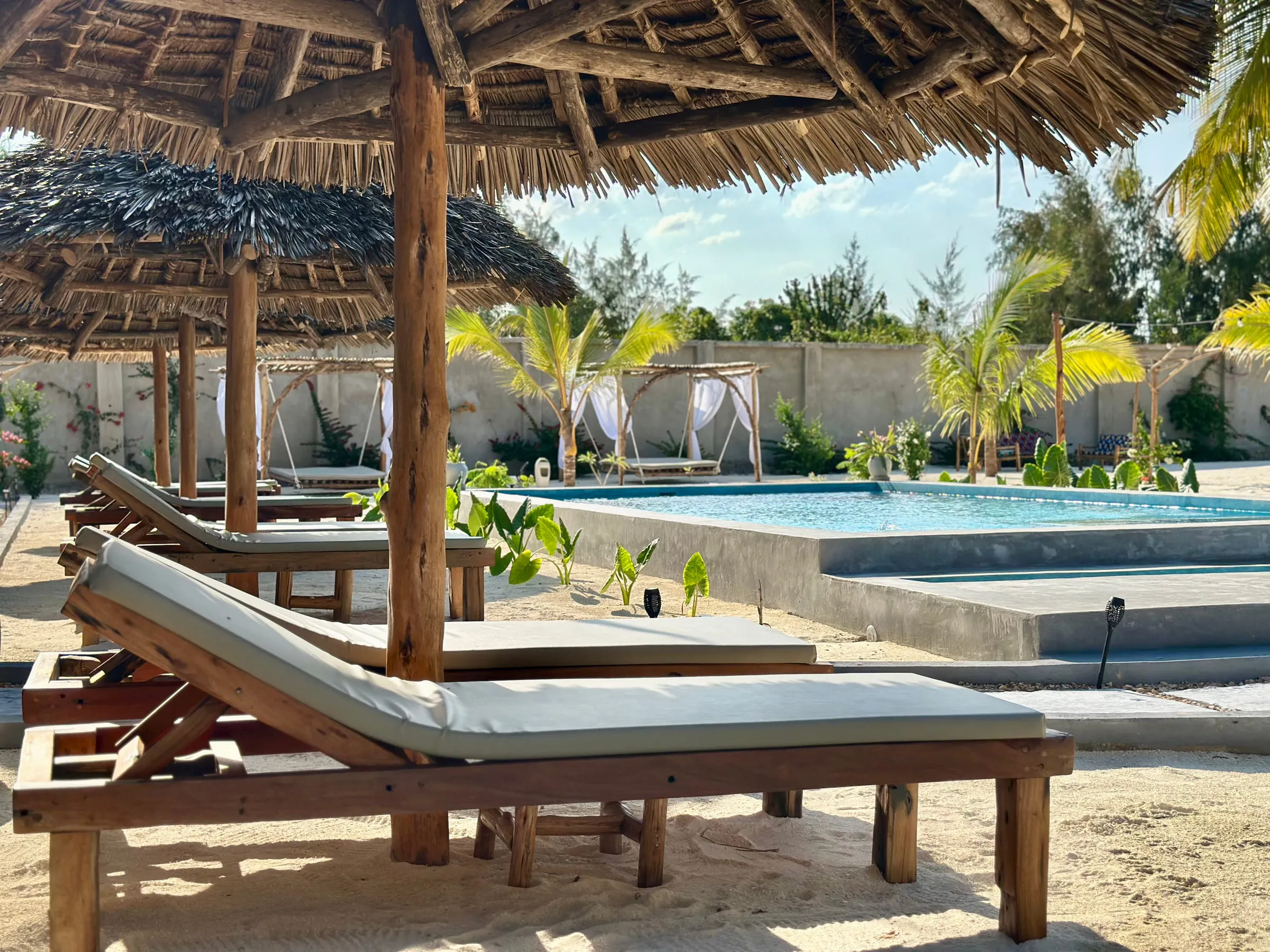 Two wooden day beds with white cushions under thatched umbrellas beside the pool