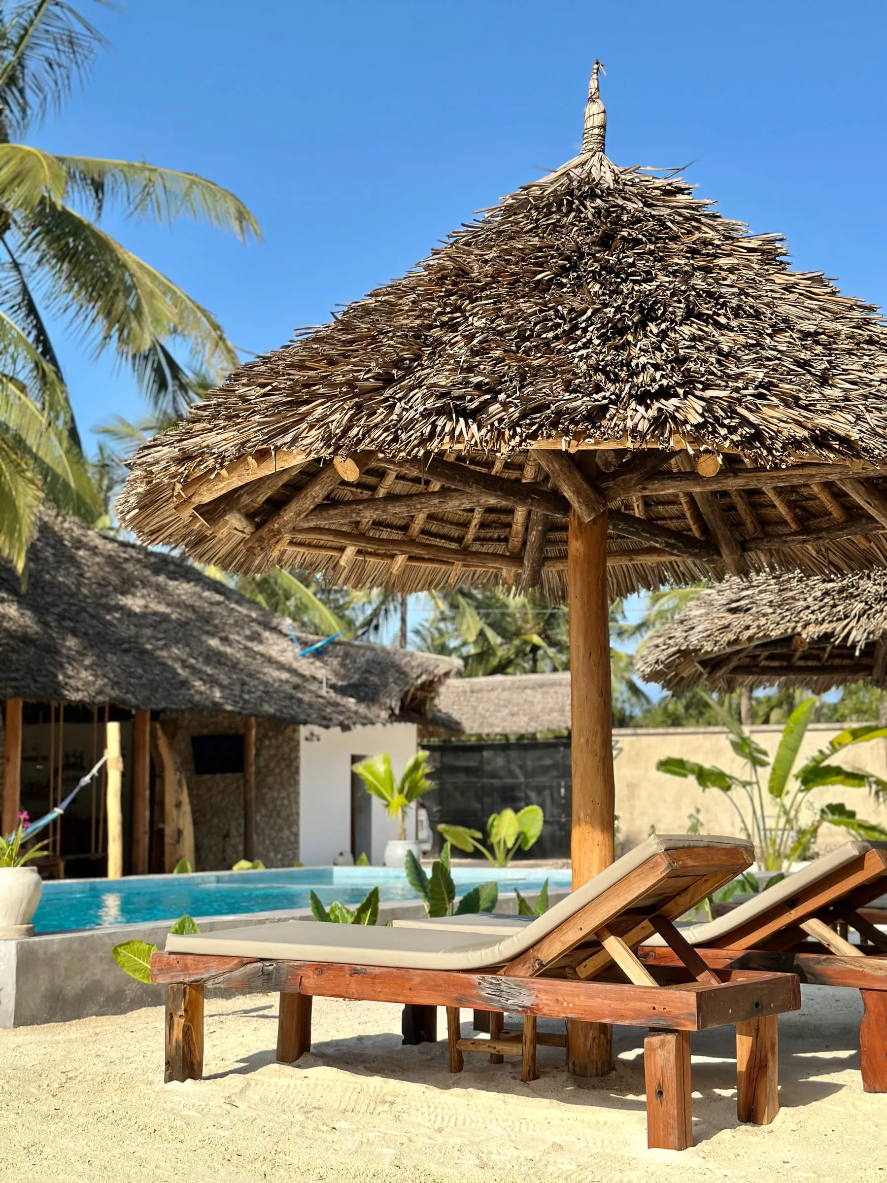 A wooden sun lounger under a tall thatched umbrella with the pool and villa behind