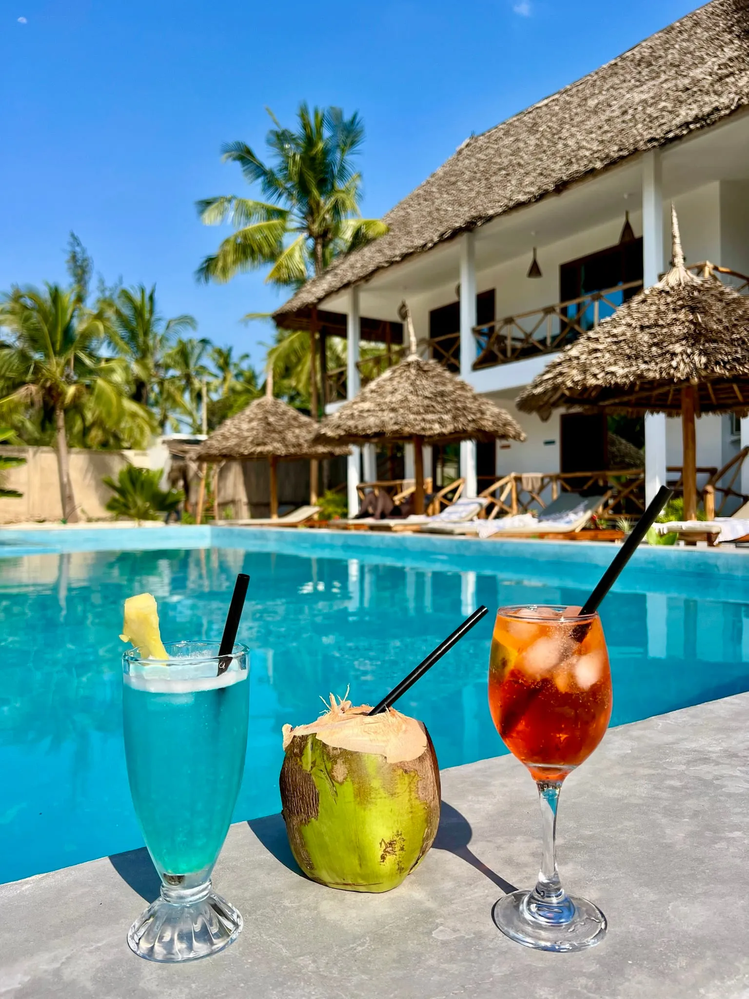 A green coconut and two coloured cocktails on the edge of the swimming pool