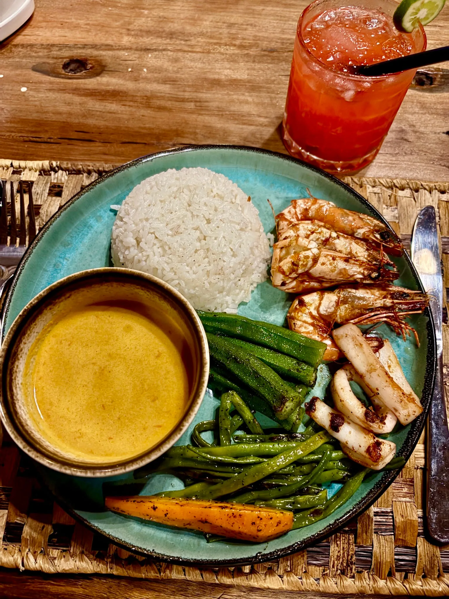 A plate of grilled fish with rice, green beans and coconut sauce beside a tall cocktail